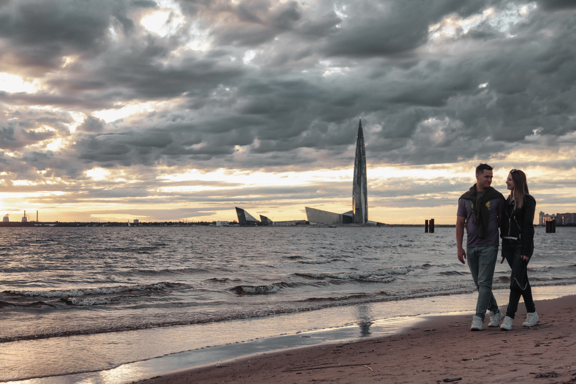 Пара гуляет по песчаному пляжу, на заднем плане виден небоскреб, Санкт-Петербург. Источник: Shutterstock