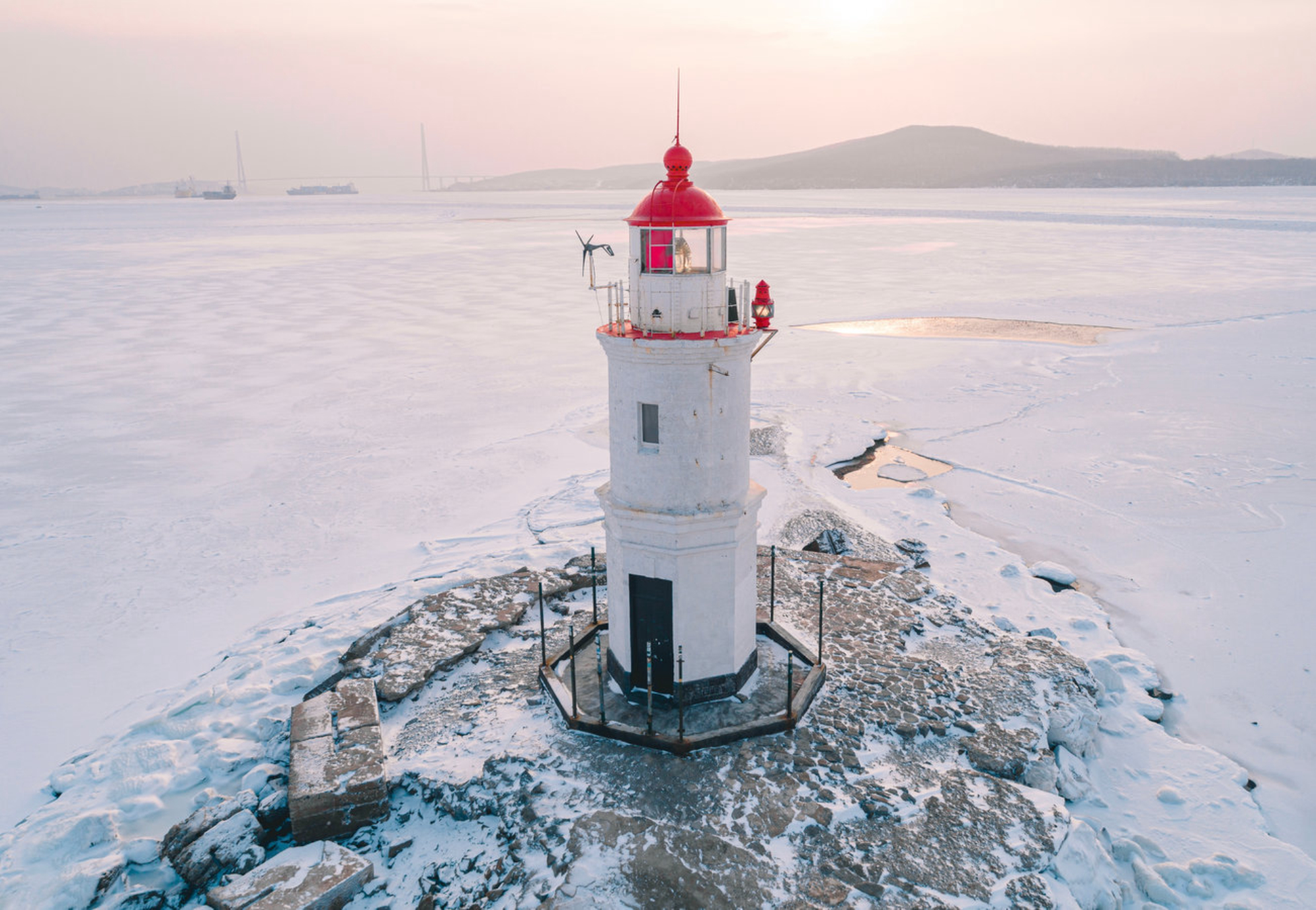 Маяк стоит на замерзшем берегу во Владивостоке во время зимнего восхода солнца с кораблями вдали. Источник: Shutterstock
