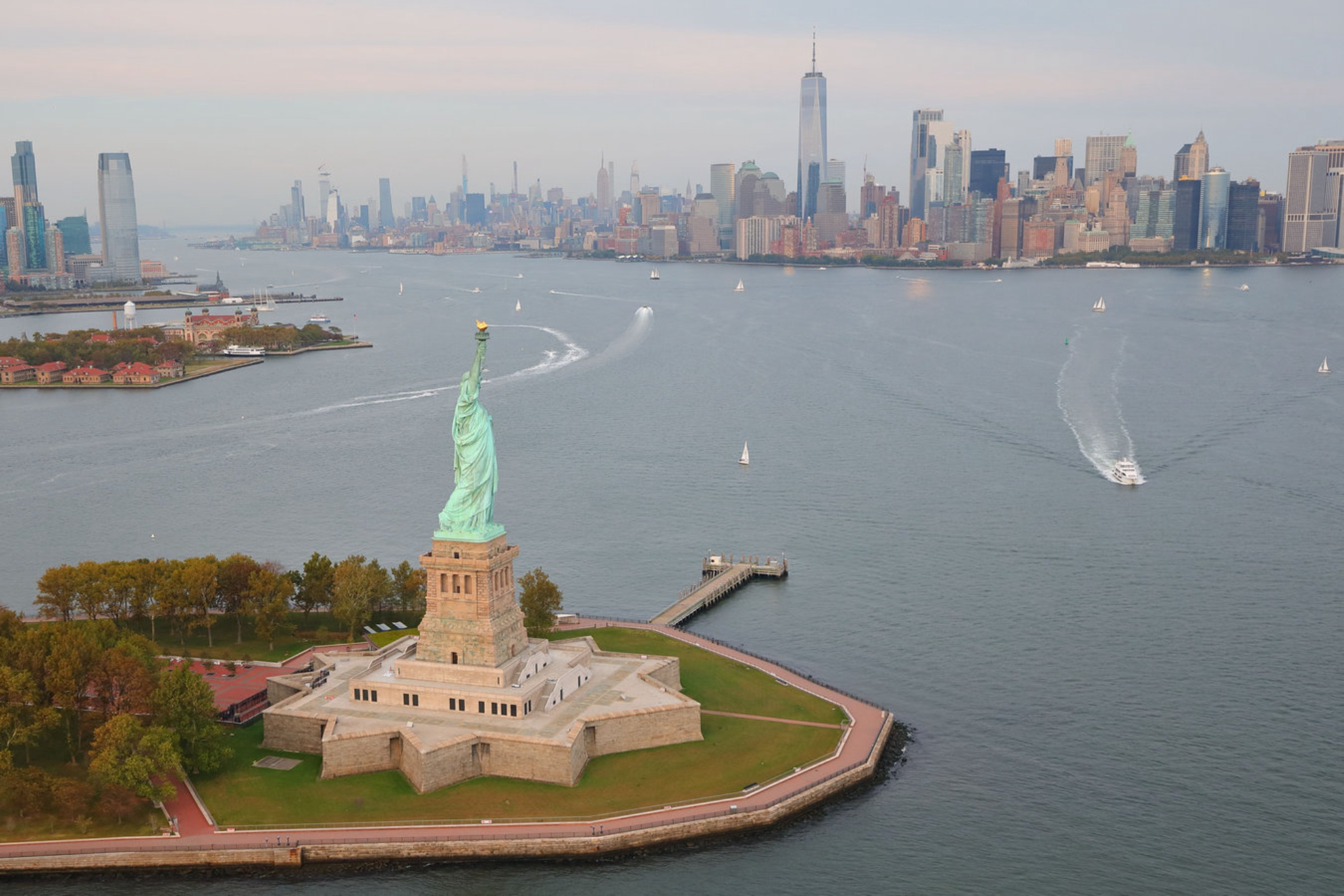 Вид с дрона на Статую Свободы на острове Свободы в окружении воды на закате в Нью-Йоркской гавани, Нью-Йорк, США Источник